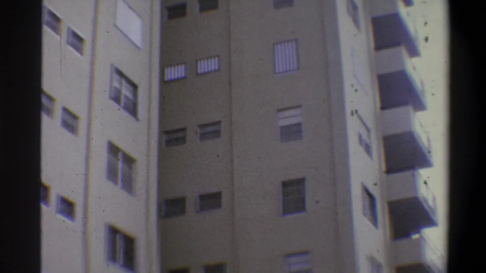 Handheld 8mm low-angle view of a tall tan concrete building with rows of louvered shutters and balconies, film grain and gate vignetting visible, circa 1973.