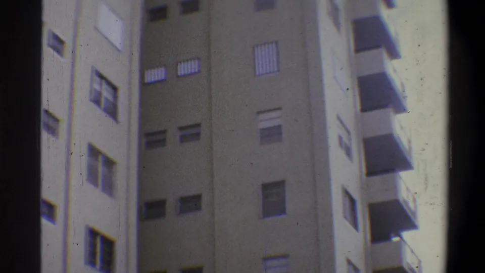 Handheld 8mm upward view of a tall tan concrete building, louvered shutters and balconies across multiple floors, gate vignetting visible, circa 1973.