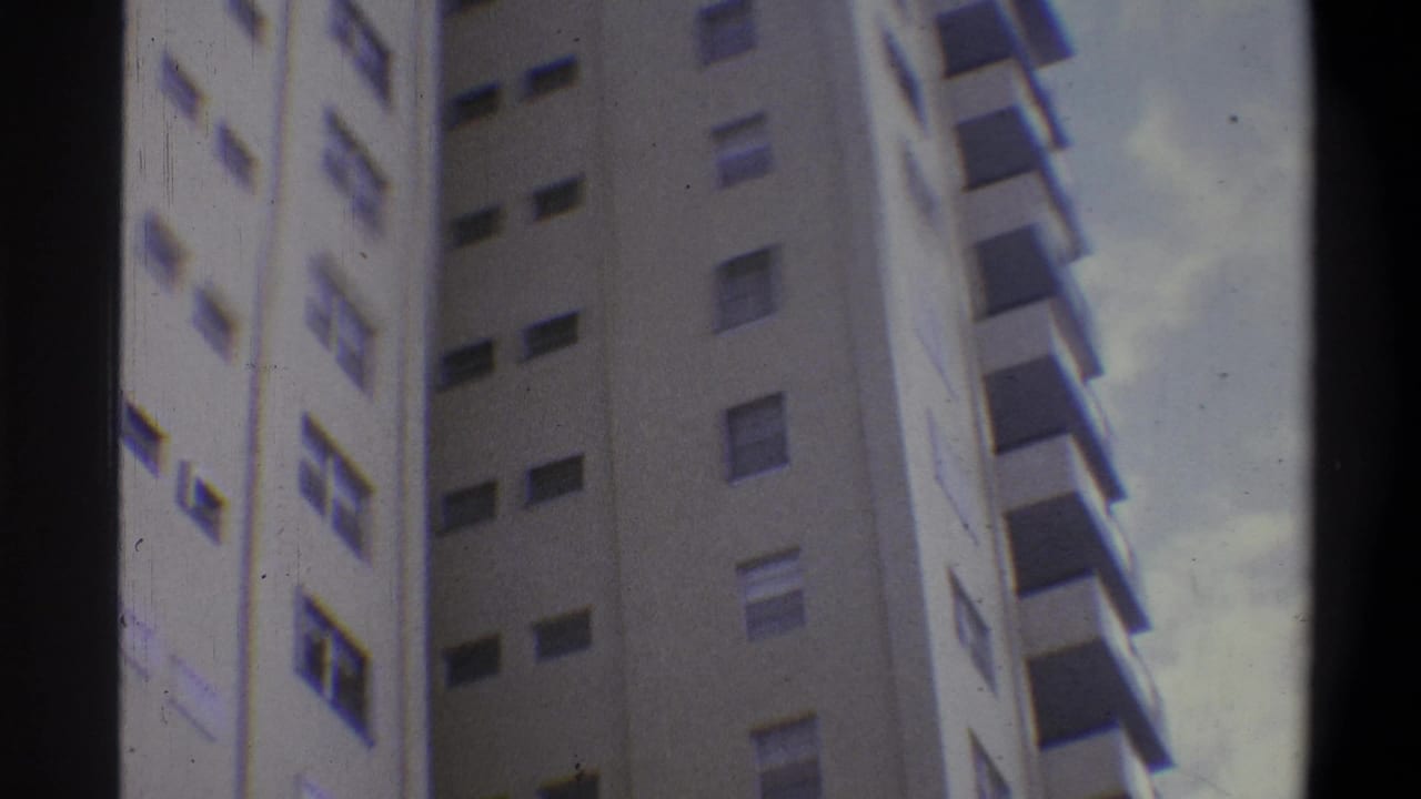 Continuing low-angle view of cream high-rise, pale sky with scattered clouds at right, possible color artifact along building edge, dark vignette at left.