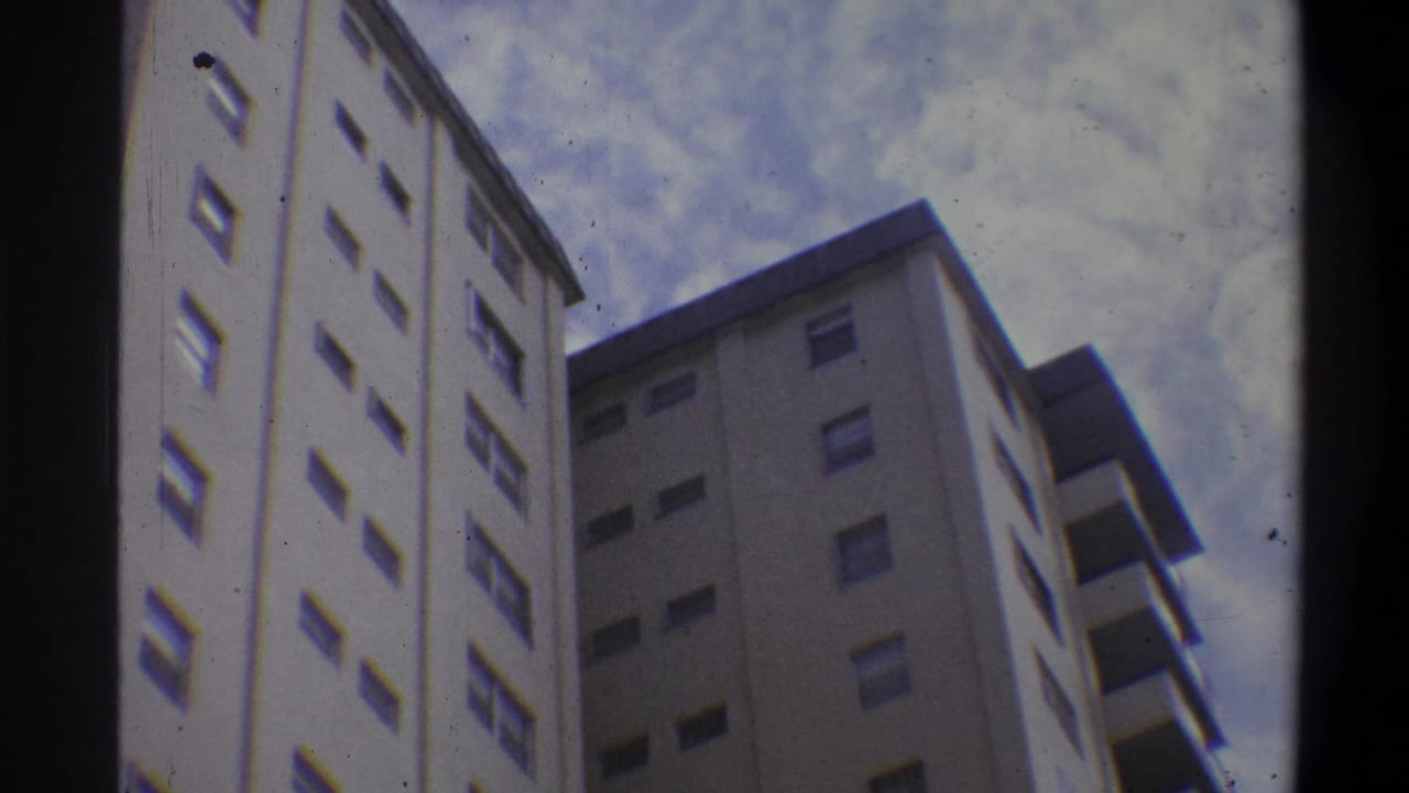 Low-angle 8mm shot of a multi-story off-white concrete apartment tower, likely 1970s, with repeating window rows and balcony overhangs against an overcast sky.