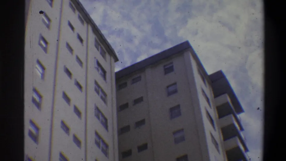 Low-angle 8mm shot of a multi-story off-white concrete apartment tower, likely 1970s, with repeating window rows and balcony overhangs against an overcast sky.