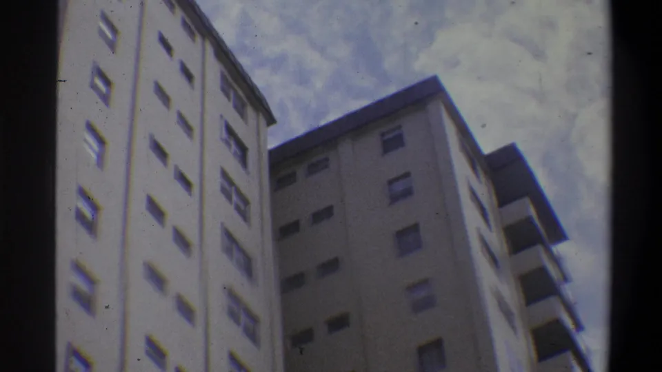 Continued 8mm low-angle shot of multi-story off-white apartment buildings with repeating window grid and balcony overhangs against an overcast sky, circa 1970s.