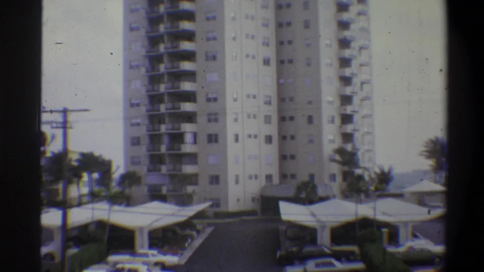Wide 8mm shot of tall white residential towers above covered parking canopies, with palm trees and utility poles in the foreground; warm-climate setting, circa 1970s.