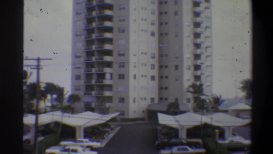 Wide 8mm view of multi-story white residential towers above covered carports, palm trees at right and utility poles visible; 1970s warm-climate setting.