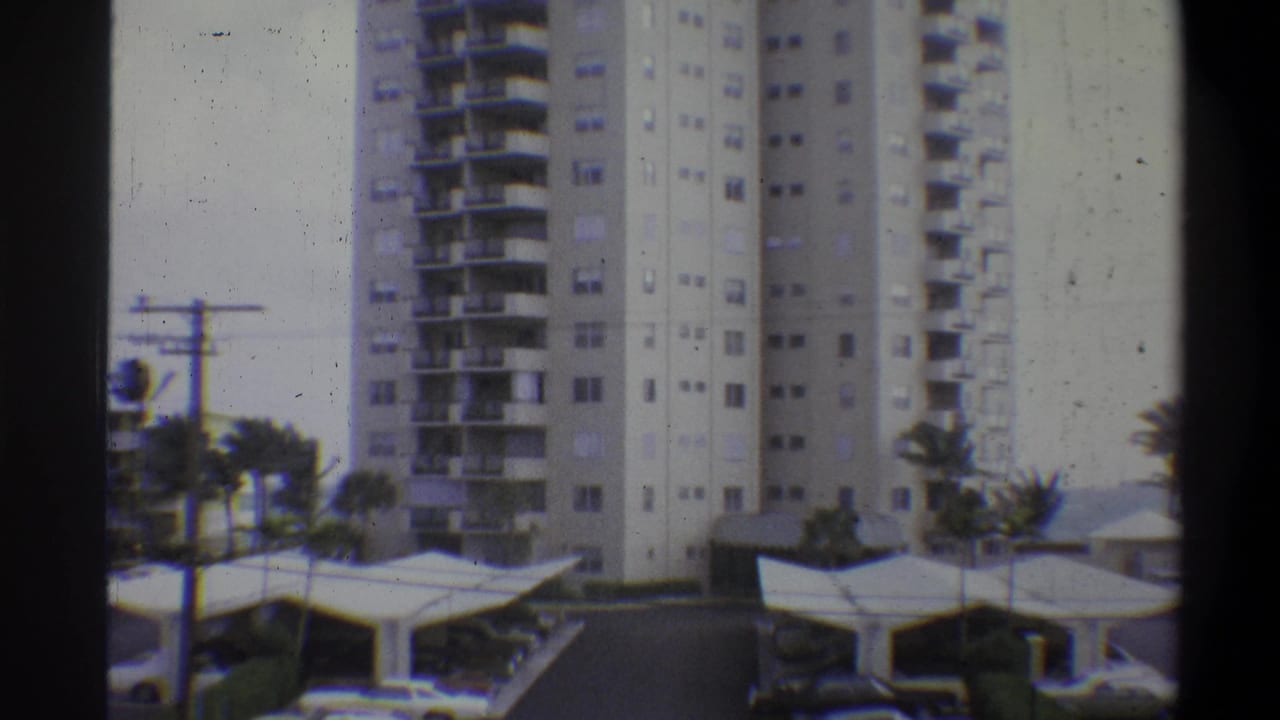 8mm color footage from 1973 showing a multi-story residential high-rise with continuous balconies, flat-canopy covered parking, and palm trees at right.