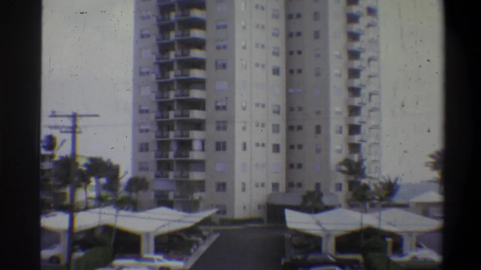 8mm color footage from 1973 showing a multi-story residential high-rise with continuous balconies, flat-canopy covered parking, and palm trees at right.