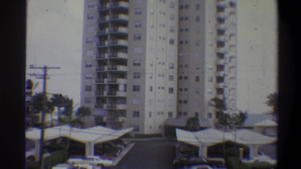 Same high-rise apartment tower with balconies; palm trees more prominent at right; flat-canopy covered parking below; overcast sky.