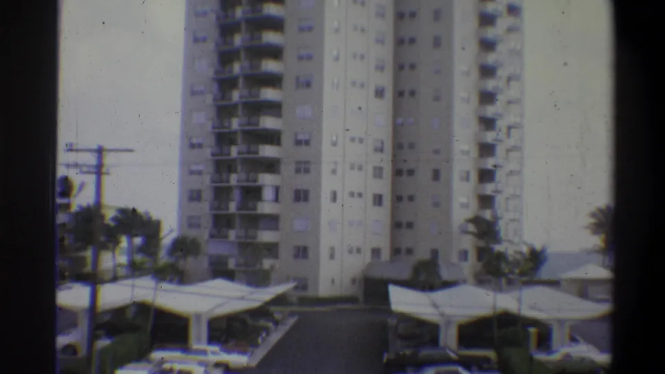 8mm color frame from 1973 showing a tall residential tower flanked by palm trees, with a mid-century flat-canopy parking structure in the foreground.