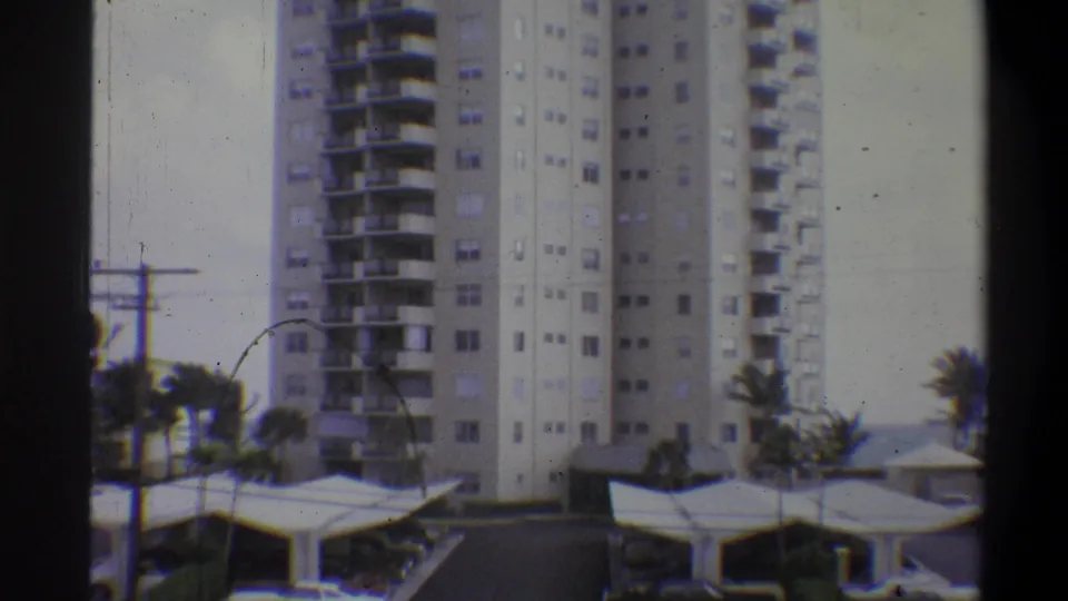 8mm color frame from 1973; slight camera shift reveals more covered parking and palm trees; a circular film scratch artifact is visible lower left.