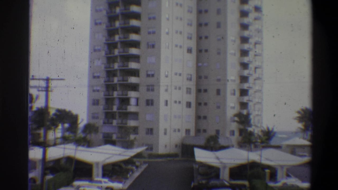 8mm home movie frame of a multi-story apartment tower with balcony grid, carport canopies and palm trees; attributed to 1973, location uncertain.