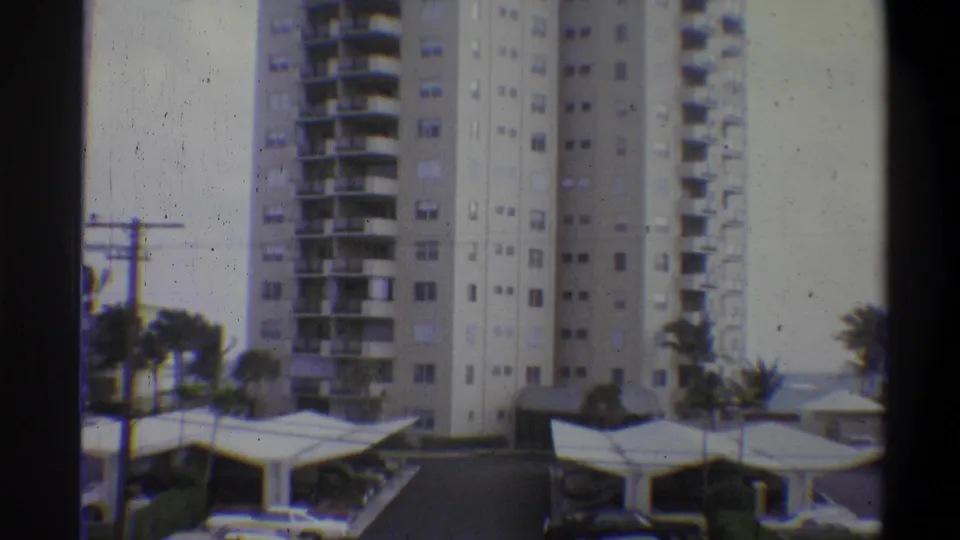 8mm home movie frame of a multi-story apartment tower with balcony grid, carport canopies and palm trees; attributed to 1973, location uncertain.