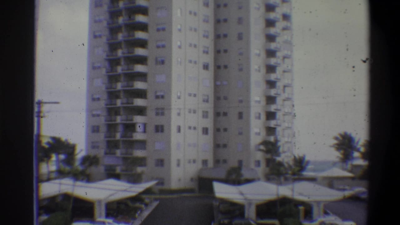 Same high-rise tower from a slightly tighter angle, carport canopies and palm trees at lower right, overcast sky, visible 8mm grain.