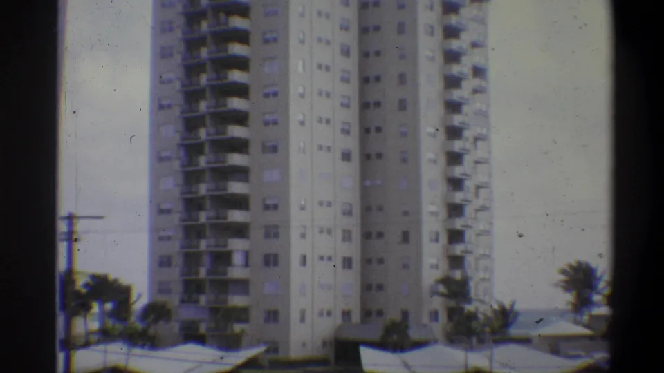 8mm home movie frame of a tall apartment tower with prominent balcony rows, palm trees at base and utility lines in the foreground; attributed to 1973.