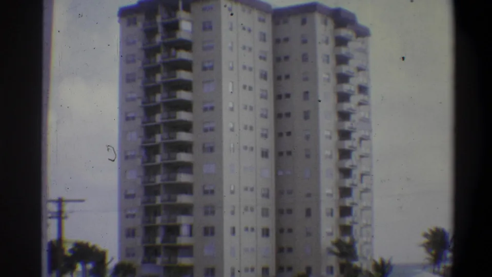 8mm film closer view of a multi-story apartment tower facade with rows of balconies and palm tree crowns at the base; attributed to 1973, location uncertain.