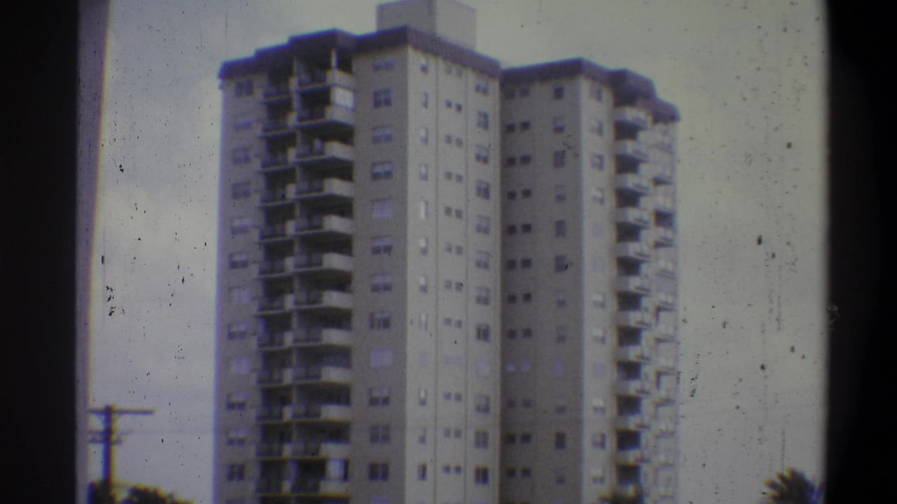 Tall apartment tower against a pale sky, slightly reframed; tree foliage at lower right; 8mm film grain and dust spots visible.