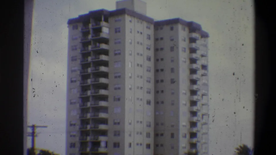 8mm home movie, Troy, New York, 1973 — the same residential tower from a slightly shifted angle, with tree foliage at lower right and film dust artifacts.