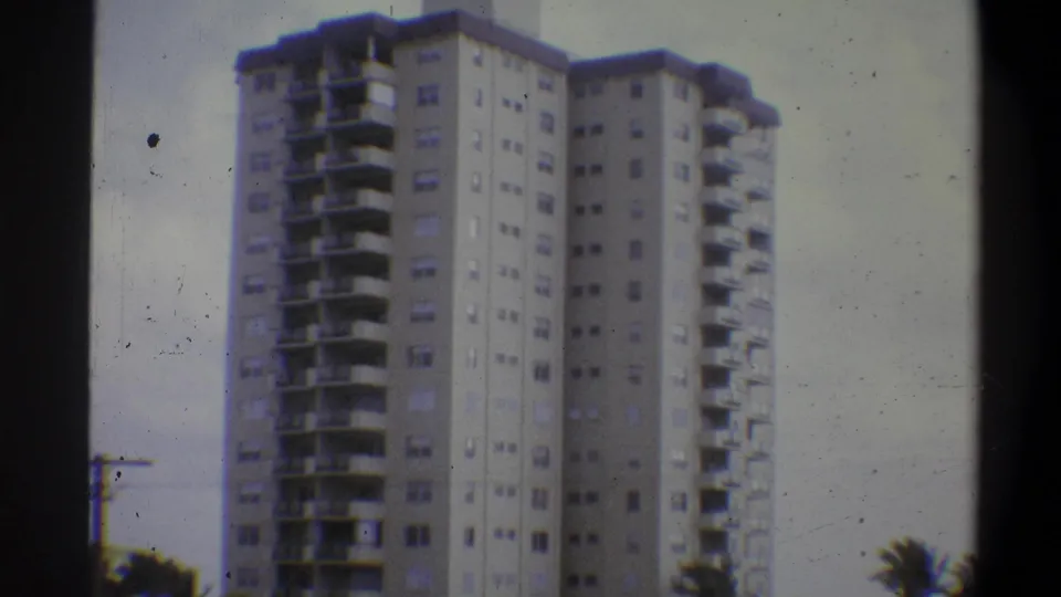 8mm home movie, Troy, New York, 1973 — residential tower upper floors against a pale sky, with tree foliage at lower right and scattered film dust artifacts.