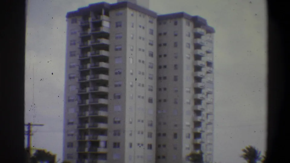 High-rise apartment building, likely Troy New York, 1973 8mm footage; low-angle view shows slab balconies, window grid, utility pole at left, trees at frame edge.