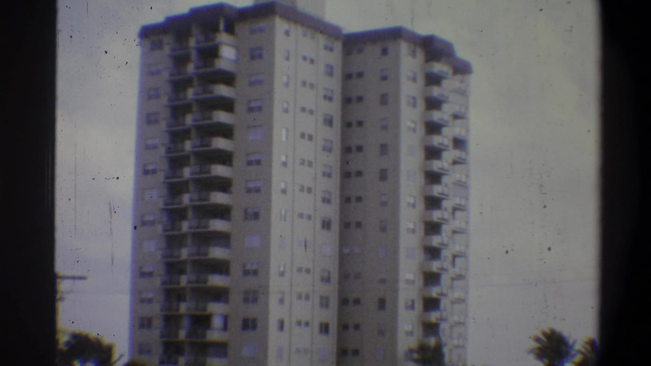 High-rise apartment tower, slightly reframed; balconied floors dominate, dark foliage at lower right, no utility pole visible, pale sky above.