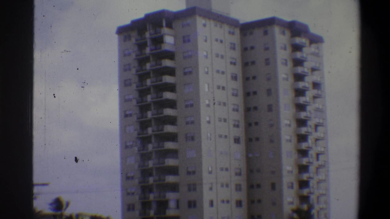 Concrete high-rise apartment tower with balconies against pale sky, 8mm film with dust artifacts.