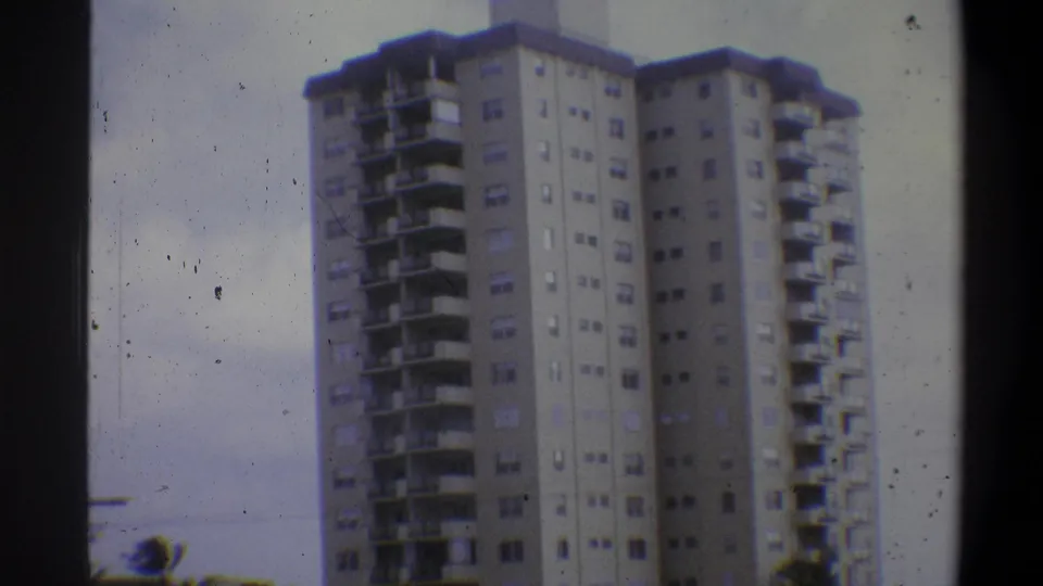 Multi-story concrete apartment building close-cropped against pale overcast sky, circa 1973, 8mm with scattered dust artifacts.