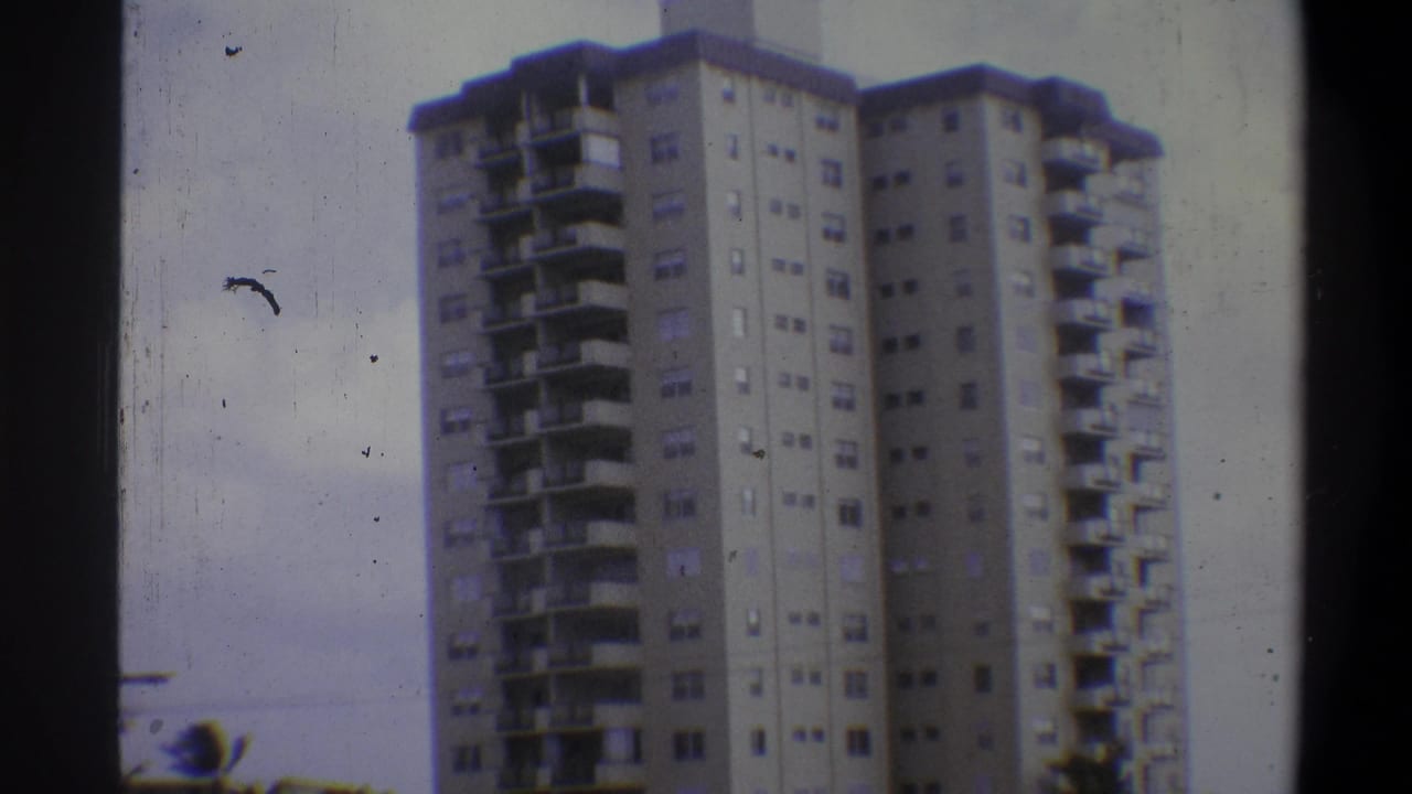 8mm home-movie frame showing the full height of a concrete apartment high-rise with stepped balconies in Troy, New York, c. 1973, under an overcast sky.