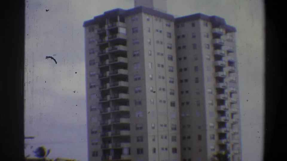 8mm home-movie frame showing the full height of a concrete apartment high-rise with stepped balconies in Troy, New York, c. 1973, under an overcast sky.