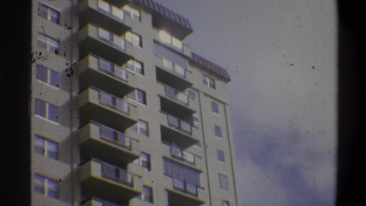 Upper floors and rooftop of a concrete apartment high-rise shot from below, emulsion debris specks visible in frame.
