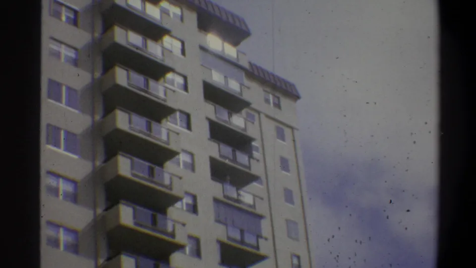 8mm home-movie frame of a modernist concrete apartment building mid-pan, balconies in sharp relief, overcast sky visible at left, Troy, New York, c. 1973.
