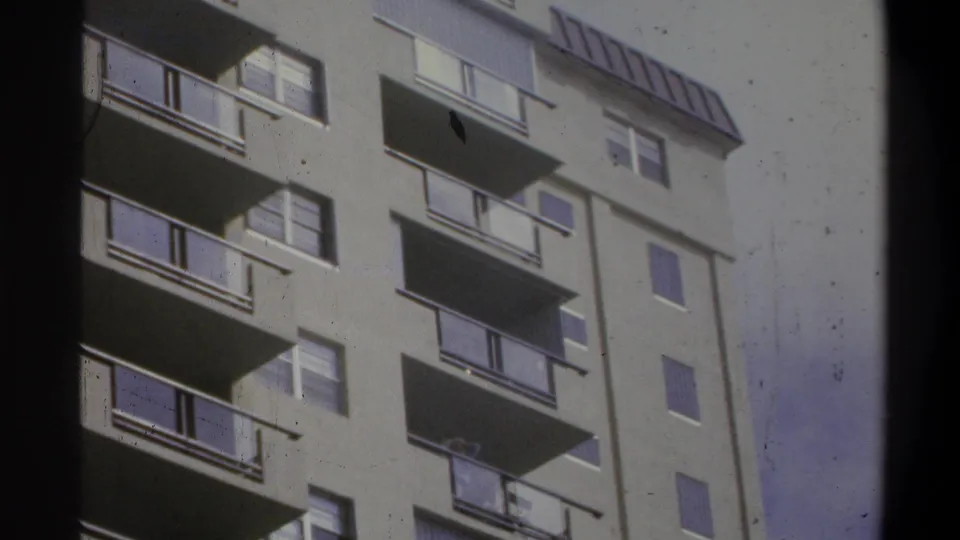 8mm home-movie close-in shot of a concrete apartment tower, Troy, New York, circa 1973, showing balconies, windows, and rooftop parapet with a visible film dust spot.