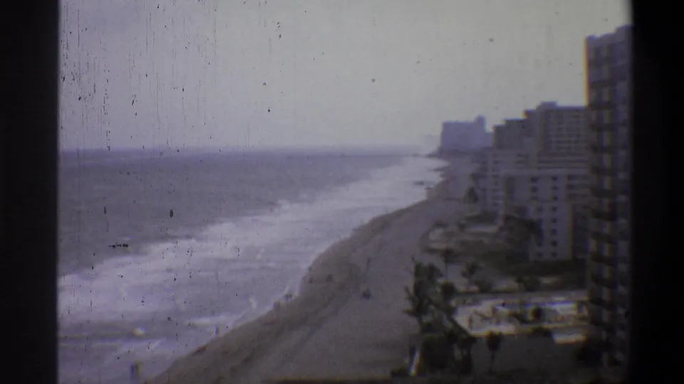 8mm home movie wide shot of a coastal beach with breaking surf and beachfront high-rises, shot from an elevated vantage point, circa 1973.