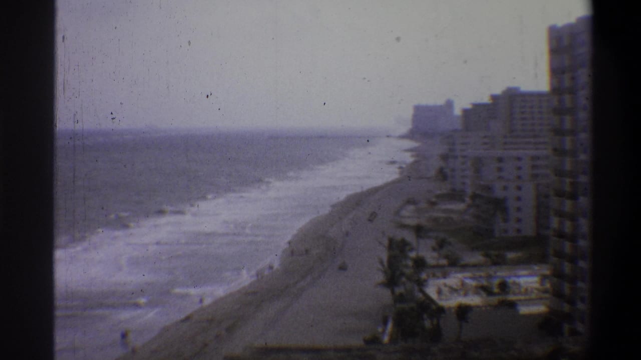 High-angle 8mm home-movie frame, likely early 1970s, showing a beachfront hotel strip, breaking waves, and palm trees under a pale overcast sky.