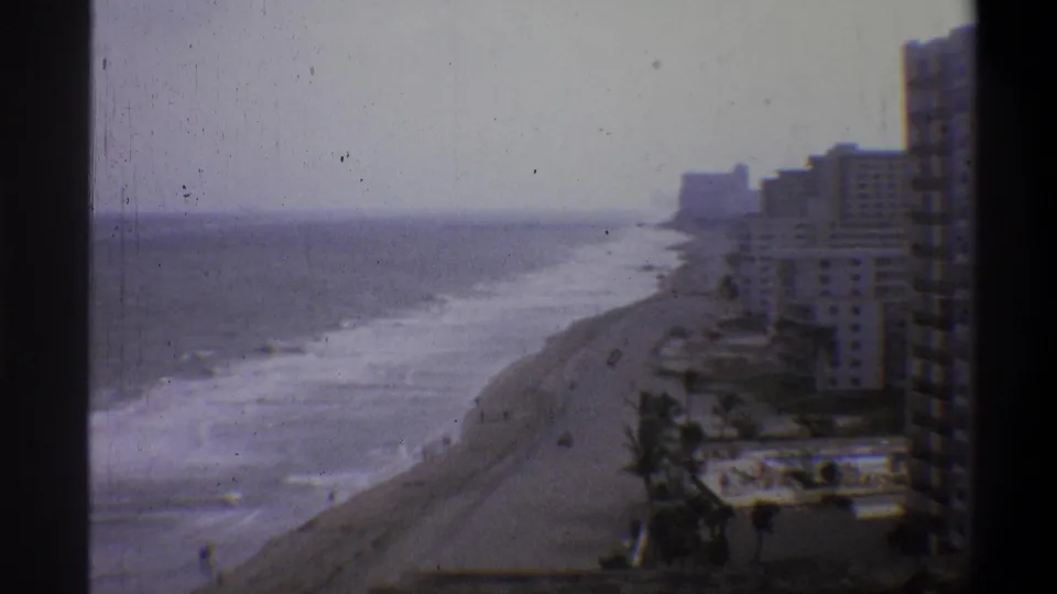 High-angle 8mm home-movie frame, likely early 1970s, showing a beachfront hotel strip, breaking waves, and palm trees under a pale overcast sky.