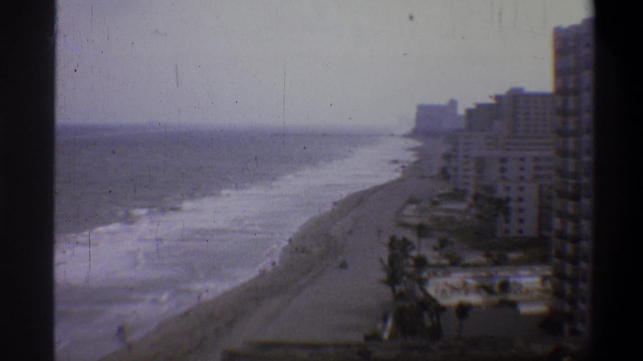 8mm home-movie frame, circa 1973, showing a beachfront hotel row, sandy shore, and breaking waves from an elevated static camera position.