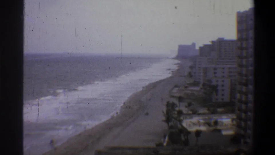 8mm home-movie frame, circa 1973, showing a beachfront hotel row, sandy shore, and breaking waves from an elevated static camera position.
