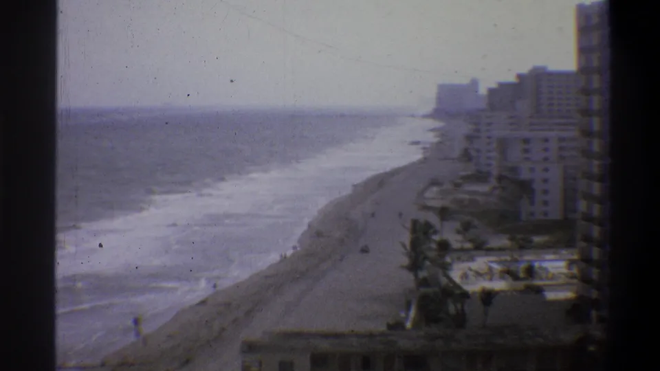 Static high-angle 8mm frame, circa 1973, showing a wide beachfront hotel strip, palm trees, and pool area under an overcast morning sky.