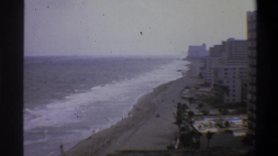 Final 8mm frame, circa 1973, showing a static wide view of a resort beach strip with hotel towers, palm trees, and breaking waves.