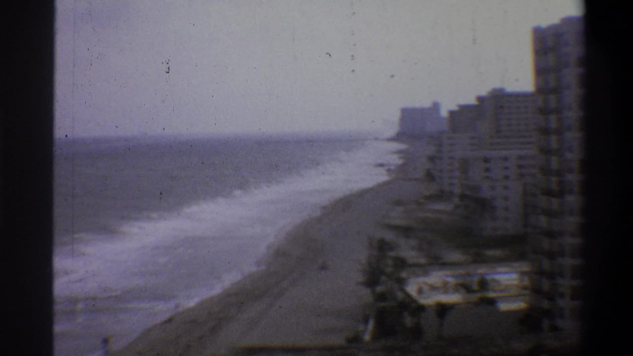 High-vantage 8mm shot of a coastal resort strip, circa 1973, showing beach, surf, and mid-century high-rise hotel row under a hazy overcast sky.