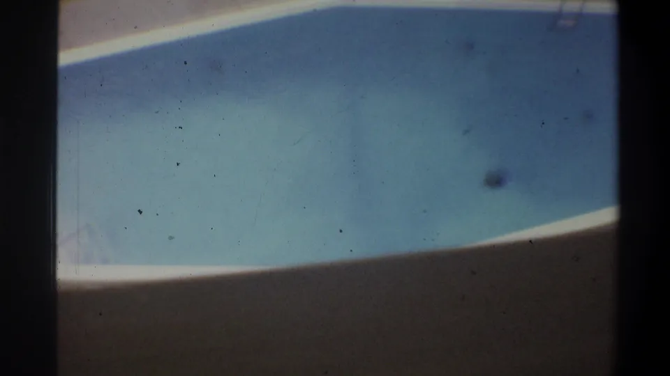 8mm overhead shot of a curved hotel swimming pool with white-tiled surround and blue water, shot from an elevated balcony, circa 1973.