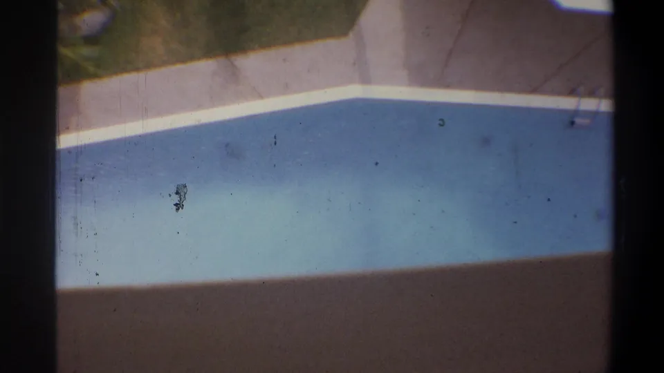 8mm overhead shot of hotel pool with white coping and surrounding deck, circa 1973; a small dark figure is visible near the pool edge — identity unclear, may be a person or film artifact.