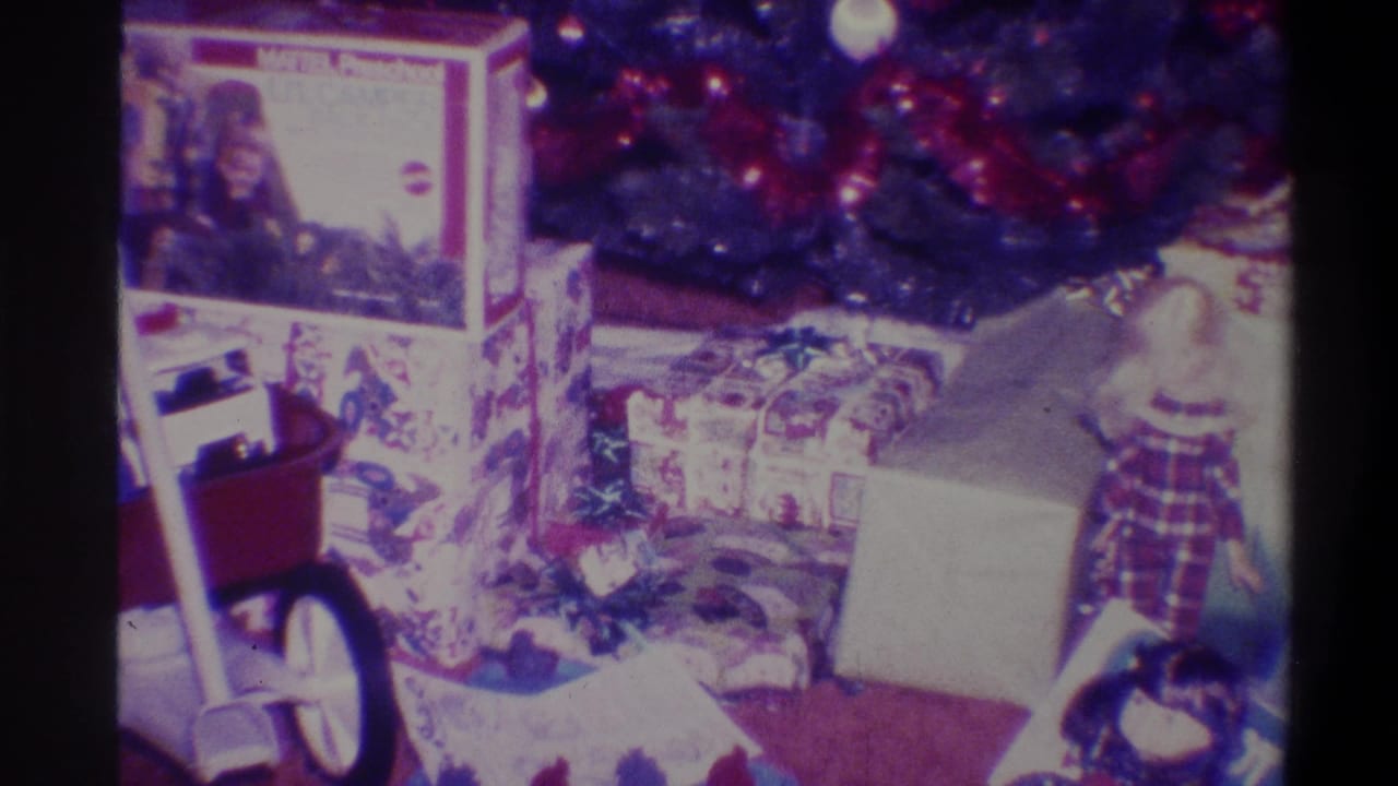 8mm home movie frame, 1973, Troy NY: stacked Christmas gifts on dark carpet beneath decorated tree, branded toy box with circular logo at upper centre.