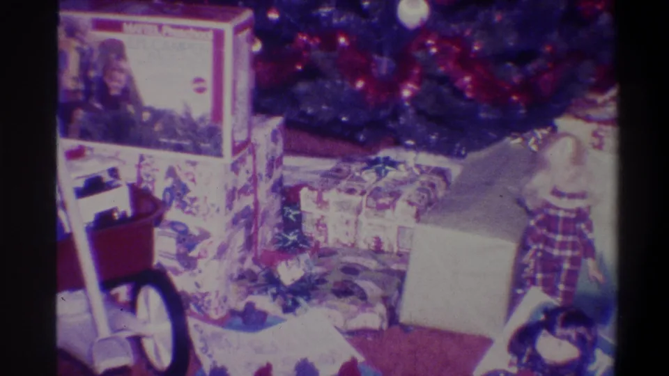 8mm home movie frame, 1973, Troy NY: stacked Christmas gifts on dark carpet beneath decorated tree, branded toy box with circular logo at upper centre.