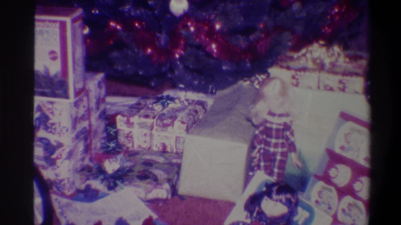Pan across Christmas gifts beneath decorated tree; branded toy box, flat wrapped package at right edge, and plaid-dressed figure visible.