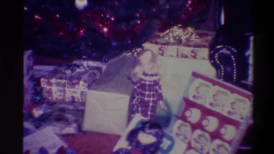 8mm home movie, 1973, Troy NY: plaid-clad figure seated among Christmas gifts on dark carpet, decorated tree above, flat boxed game item in foreground.