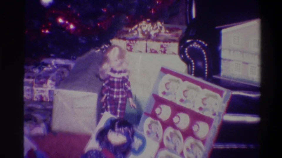 8mm home movie, 1973, Troy NY: plaid-dressed figure among Christmas presents, flat box with circular graphic design in foreground, window light at right.