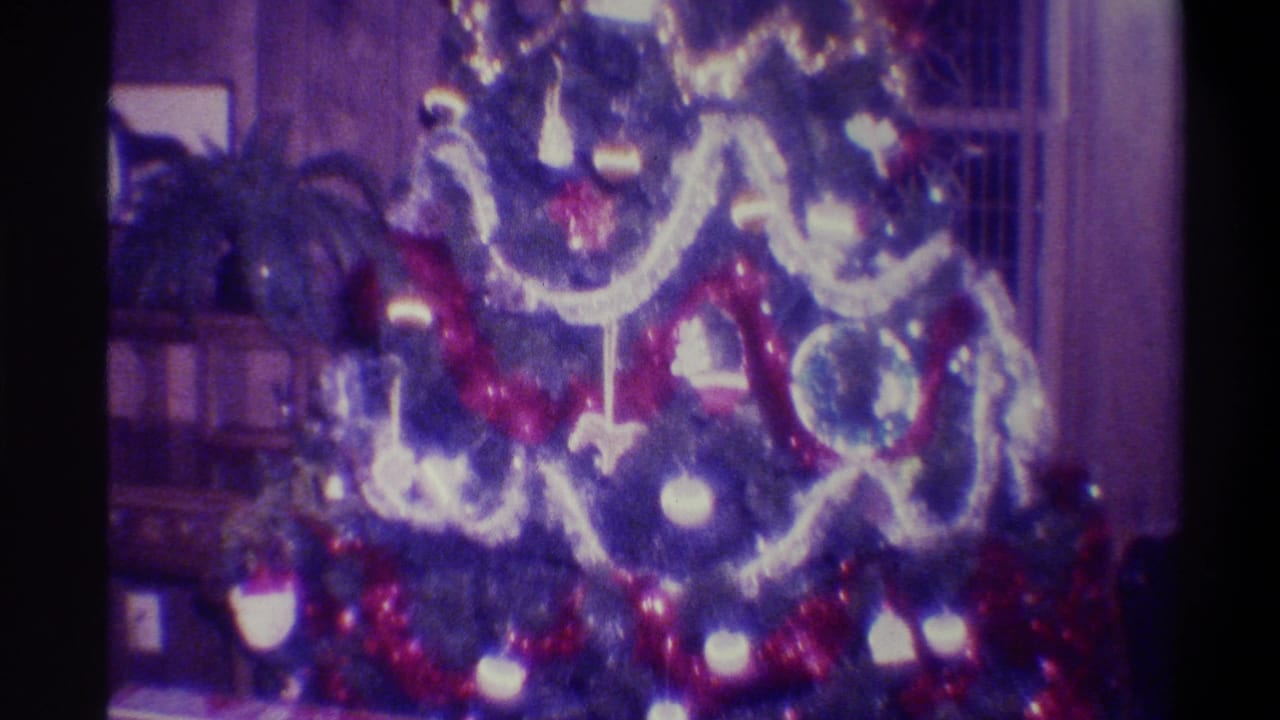 Upper portion of Christmas tree with red tinsel, silver garland, and round glass ornaments; shelving and houseplants visible in background; purple-shifted 8mm film.