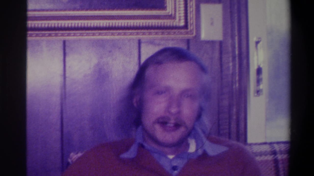 1973 home movie frame of a man in a red sweater speaking to the camera in a wood-paneled living room in Troy, New York, with plaid couch edge and framed artwork visible.