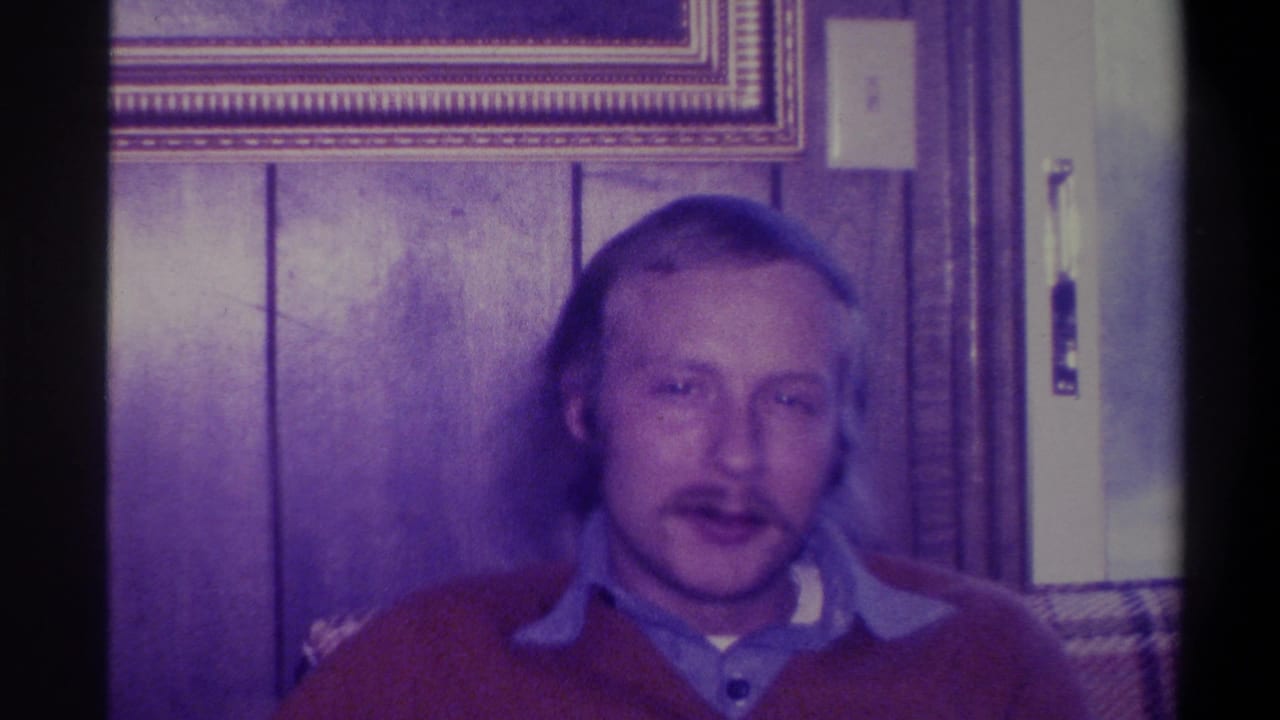 Same man in red sweater mid-speech facing camera, wood paneling and door hardware visible behind him, purple-cast Super 8 film.