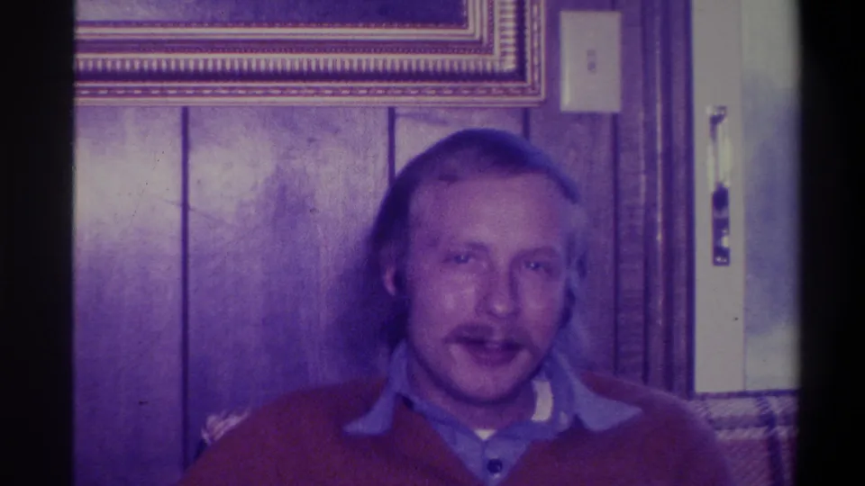1973 home movie frame of the same man in a red sweater, now slightly closer to the camera, still addressing it directly in a wood-paneled room.