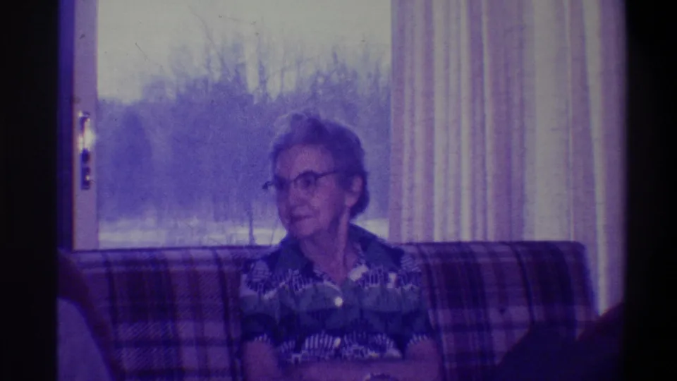 1973 home movie frame of an elderly woman with glasses and a patterned blouse, seated on a plaid couch in front of a curtained window with bare trees outside.